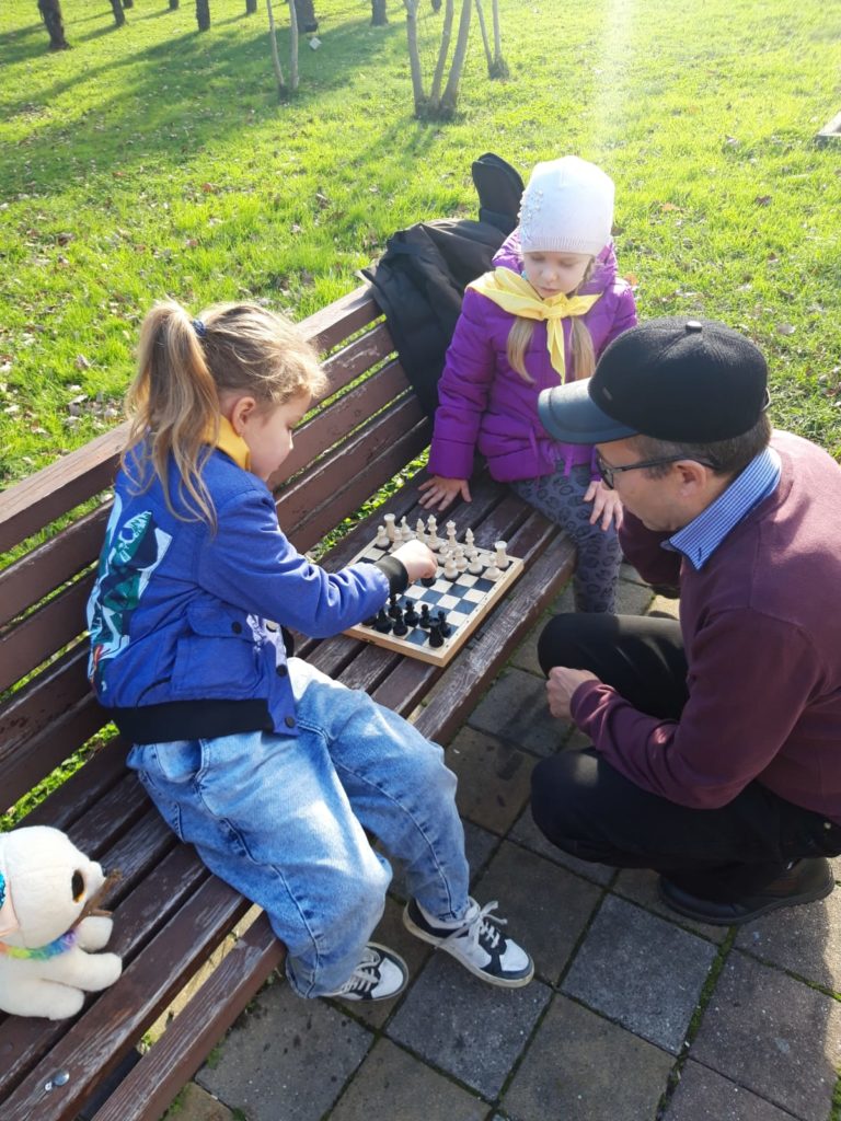 Two girls playing chess with grandpa on a bench in a park
